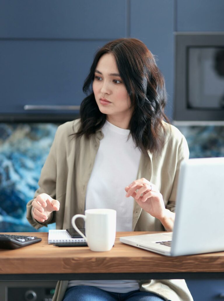 Focused young woman calculating budget while sitting with laptop and coffee at home.